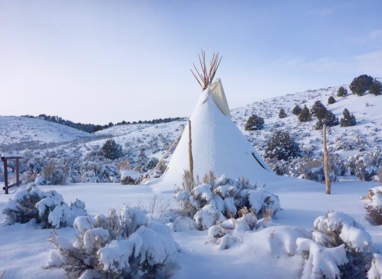A snow-covered tipi sits in a hilly landscape dotted with trees and bushes against a blue sky. Partially obscured.