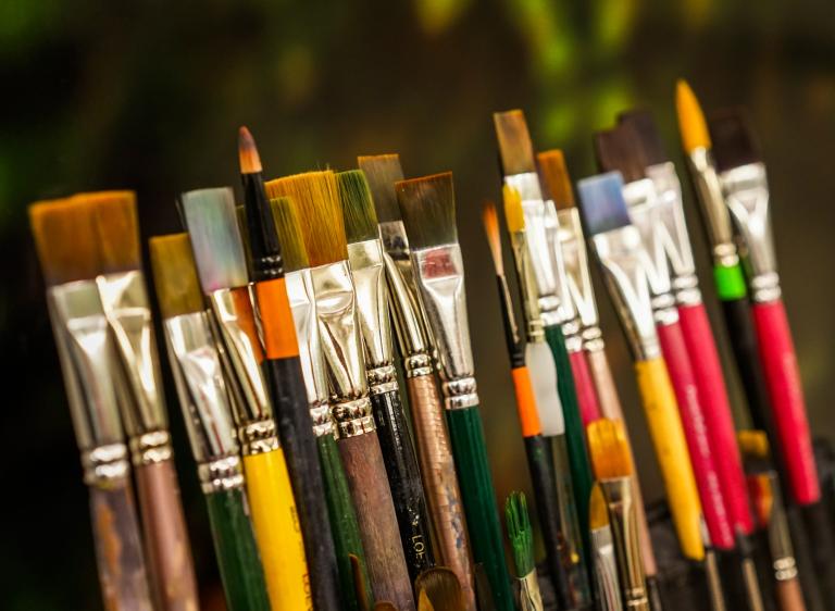 Various sizes of paint brushes standing in a paint-brush holder against a blurred green backdrop. Partially obscured.