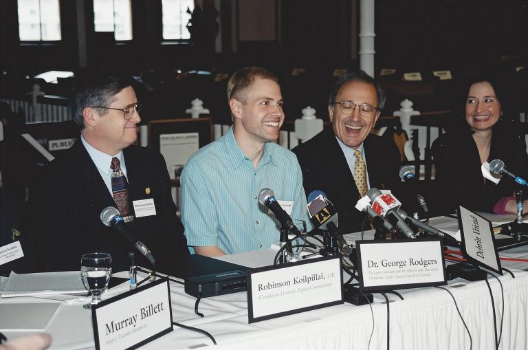 A man wearing a light blue shirt sits at a long table in front of a cluster of microphones, smiling. Seated on both sides of him are other people dressed in suits and facing microphones, also smiling. The name placards along the front of the table read, from left to right, “Murray Billet”, “Robinson Koilpillai”, and “Dr George Rodgers”, with too-blurry-to-read affiliations below the names. Another placard, knocked on its side, reads “Delwin Vriend” without an affiliation. Partially obscured.