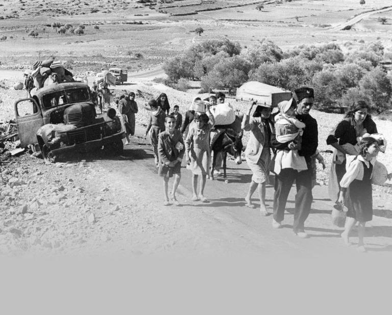 A black-and-white photo of a group of adults and children carrying belongings, walking down an unpaved desert road past a broken-down truck. Partially obscured.