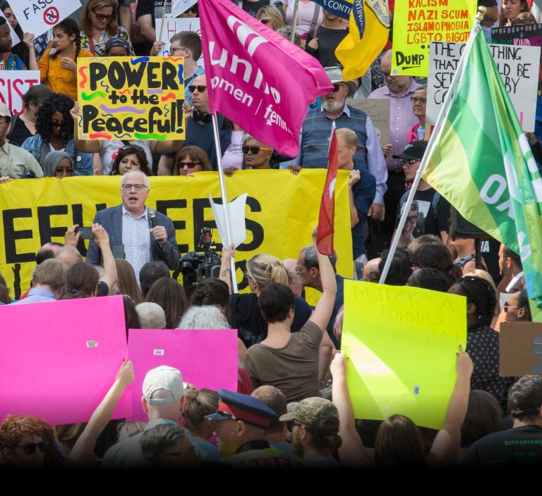  A white-haired white man with glasses and wearing a blazer is speaking and holding a microphone. Around him is a crowd of people holding cameras, protest signs and a banner. Partially obscured.