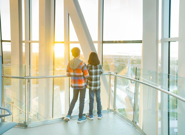Two young children stand in a tower with their backs toward the camera as they stare out at a horizon overlooking a cityscape. Partially obscured.
