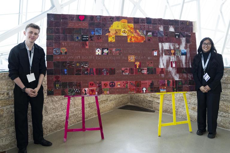 Students standing next to their large artwork displayed on easels. Partially obscured.