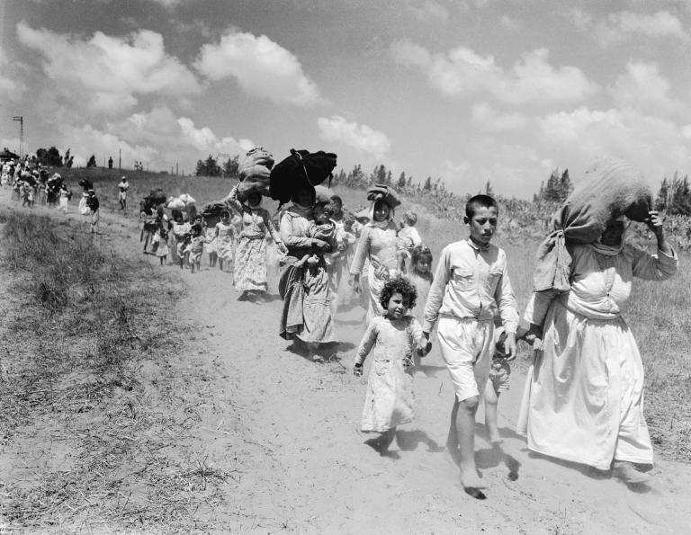 Women and children carrying bundles on their heads walk along a dirt road. Partially obscured.