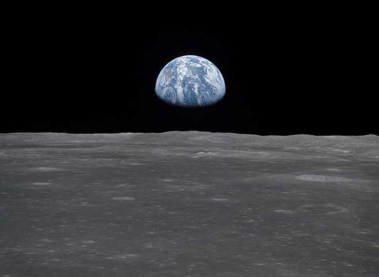 View of earth rising above the lunar horizon, showing the blue and white planet against the dark backdrop of space as seen from the moon’s surface. Partially obscured.