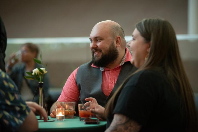 A man and woman sit together at a dinner table, smiling warmly as they listen to other guests. The man wears a salmon shirt with a gray vest, and there are small candles and drinks on the table, creating a cozy intimate atmosphere. Partially obscured.