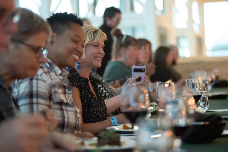 A group of people sit at a long dining table, laughing and enjoying a meal together. The focus is on two people smiling brightly - one wearing a plaid shirt and the other in a black patterned top, with glasses and plates arranged neatly in front of them. Partially obscured.