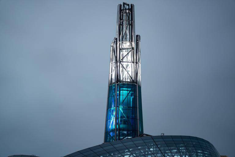 A tall, illuminated glass-and-steel spire rises above the curved patterned roof of the Canadian Museum for Human Rights, glowing in blue and white light against a soft grey sky. Partially obscured.