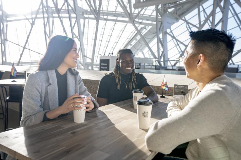 Three people are seated at a table. They're all smiling with coffees in front of them. There is a small progress pride flag on their table and at the empty table behind them. The sun is shining bright through the Museum windows. Partially obscured.
