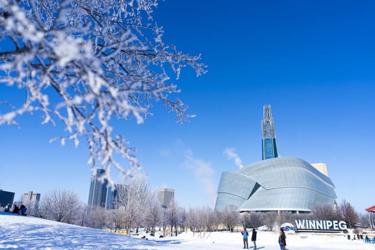 An outdoor winter scene with an exterior view of the Canadian Museum for Human Rights, other buildings in the distance and trees covered in frost. Partially obscured.