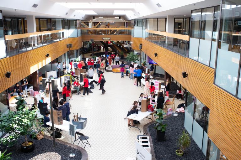 Many people gathered in an open indoor space with tables and posters set up on them. Partially obscured.