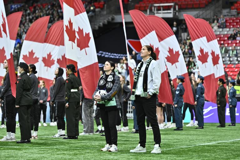 People stand on a stadium field during a pre-game ceremony as large Canadian flags are held behind them, filling the background with red and white. Partially obscured.