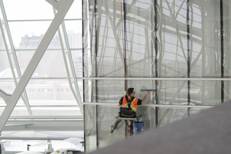 Male worker wearing safety vest suspended from ropes cleaning windows from a high height. Partially obscured.