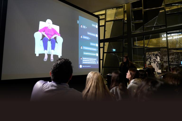 Audience members view a large projection screen displaying an elderly woman in a purple top and blue pants sitting in a white armchair, alongside an interactive message interface. The presentation takes place in a modern space with geometric glass architecture, and a presenter stands to the right of the screen. Partially obscured.