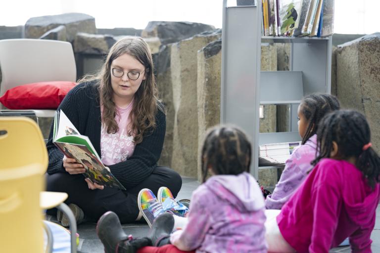 An adult sitting on the floor reading a book to three children sitting in front of them. Partially obscured.