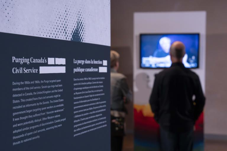 An exhibit panel with the text “Purging Canada’s Civil Service” is on the left. Two people in the background on the right watch a video. 