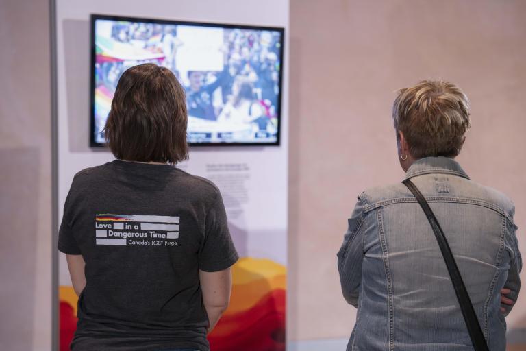 Two people stand with their backs to us and watch a video. The wall holding the video monitor features rainbow colours.