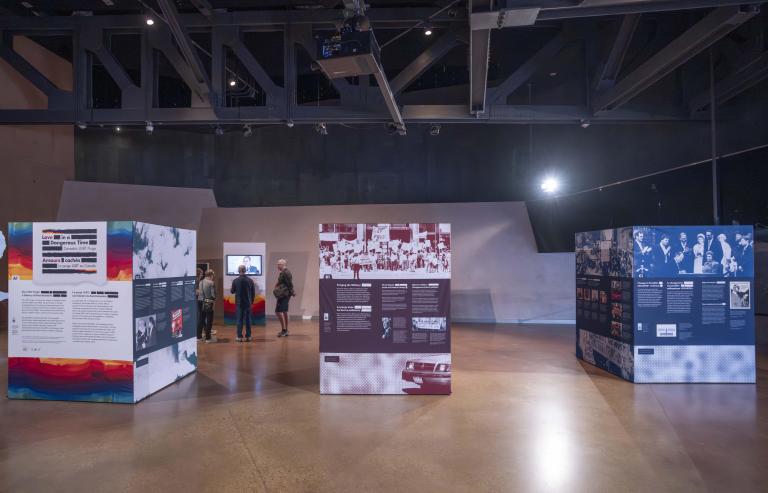 Three large cubes, taller than a person and arranged side-by-side, are covered with images and text. These cubes are the pop-up exhibit Love in a Dangerous Time.