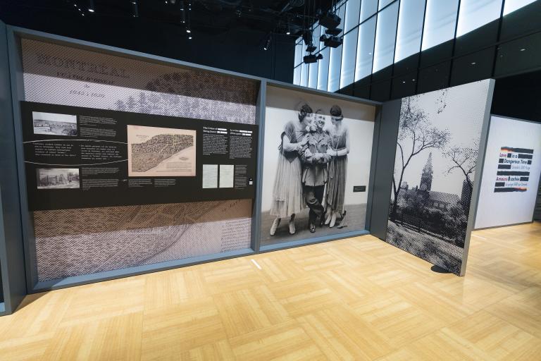 A large exhibit panel with text and historical images is on the left. On the right is a large black-and-white photograph from the 1910s of a man in a military uniform flanked by two women in dresses. A photograph of a large building can be seen further to the right.