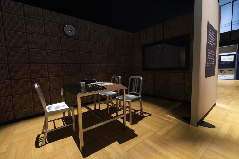 Three metal chairs and a metal table covered with binders and a tape recorder are in the middle of a darkened space. A clock hangs on the wall above the table. A mirrored surface is to the right of the table.