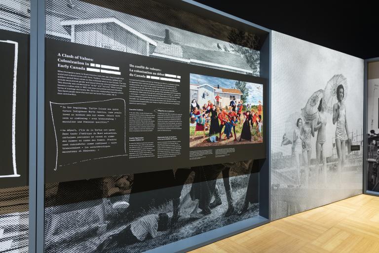 A large exhibit panel on the left features the words “A Clash of Values: Colonization in Early Canada” and a colourful painting. On the right, there is a large black-and-white photograph of people parading in single file in 1940s women’s swimsuits.