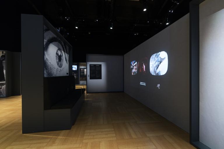An exhibition panel on the left has a picture of a large eye on it. On the right, three small videos are projected on to the wall. The space is dark.