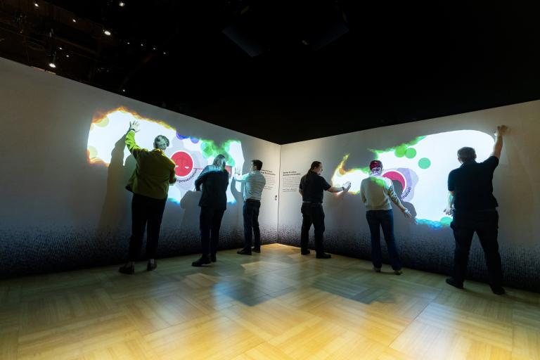 A group of people wave their arms in front of a darkened wall. Colours, shapes, and words appear on the wall where their hands are.