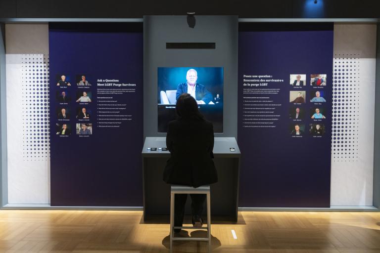 A person with their back to us watches a video. Small portraits of people flank the video monitor on the left and right.