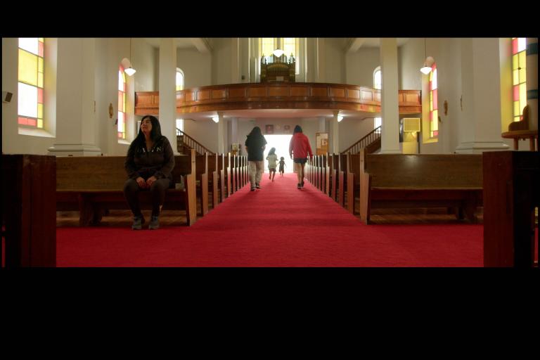 A church interior with a long red-carpeted aisle between wooden pews. Sunlight filters through stained-glass windows as several children run toward the bright entrance at the far end while an Indigenous woman sits alone in a pew to the left. Partially obscured.