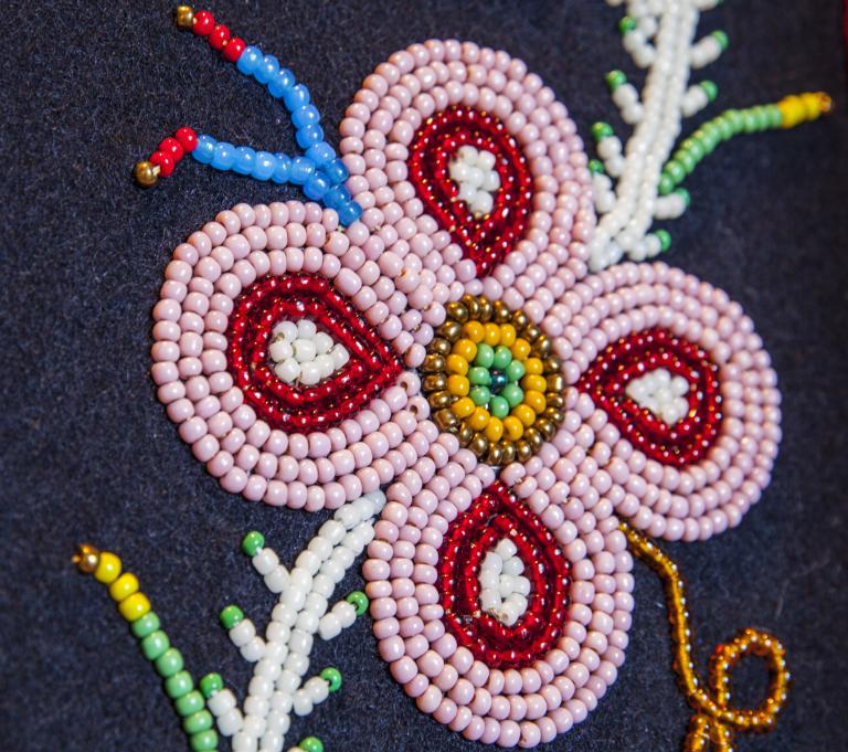 Close-up of a beaded floral design on dark fabric, featuring a pink four petal flower with red and white details, a yellow and green beaded center, and surrounding beadwork resembling leaves and stems. Partially obscured.