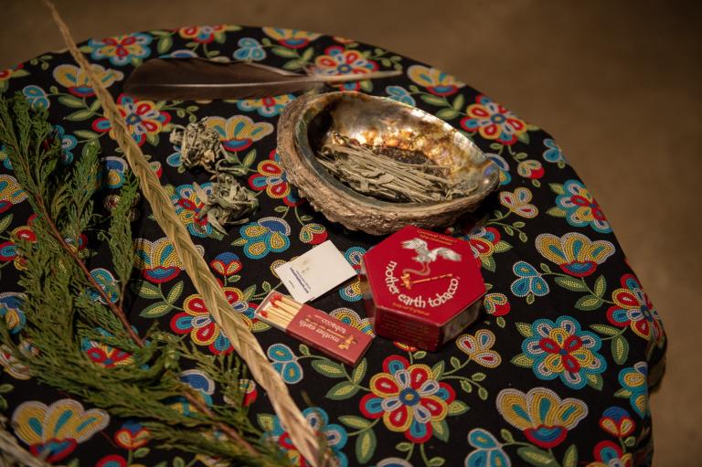Smudging items arranged on a round table covered with a floral cloth, including an abalone shell holding dried sage, a feather, braided sweetgrass, cedar branches, and a small box of matches. Partially obscured.