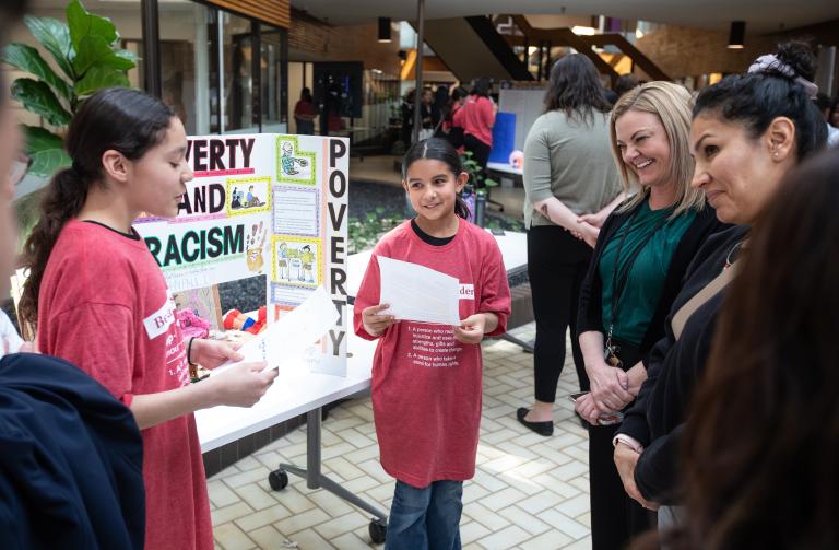 Two students standing in front of their project displayed on a table, presenting their work to visitors. Partially obscured.