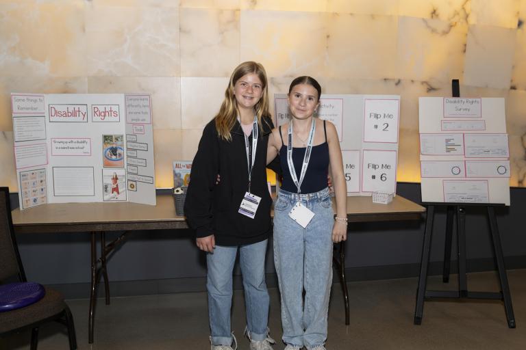 Two students standing in front of a table and an easel that are displaying presentations on poster boards. 
