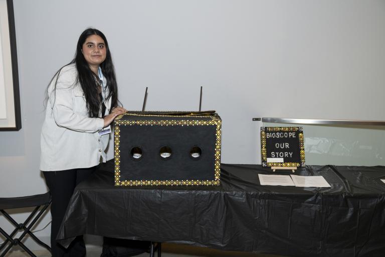 A young woman stands next to a box with an ornamental painted trim. The box has three holes that give a view into the box. 