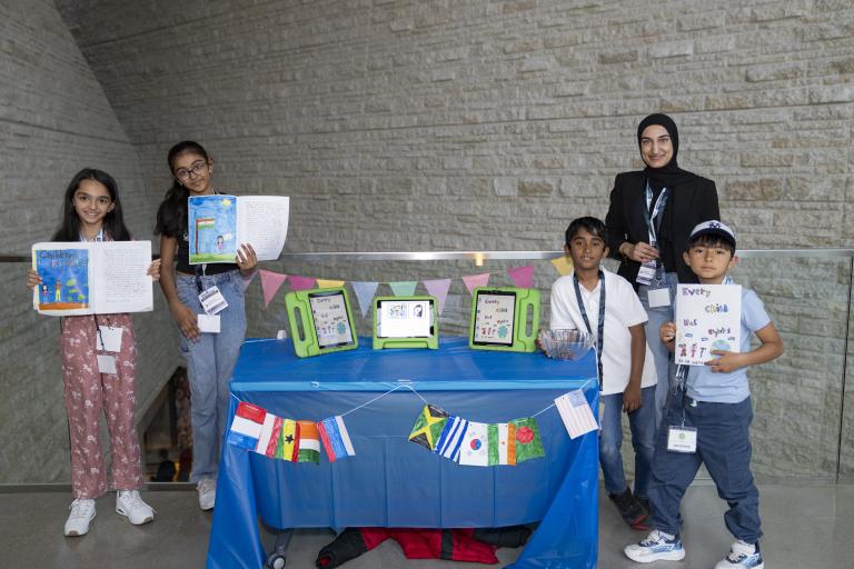 Four students and an adult stand next to a table with flags from countries around the world. Three of them are holding up an open book.
