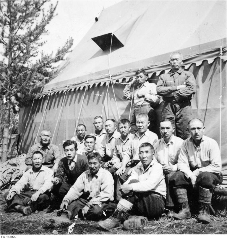 A group of men pose for a picture with a large tent and a tree in the background. Partially obscured.