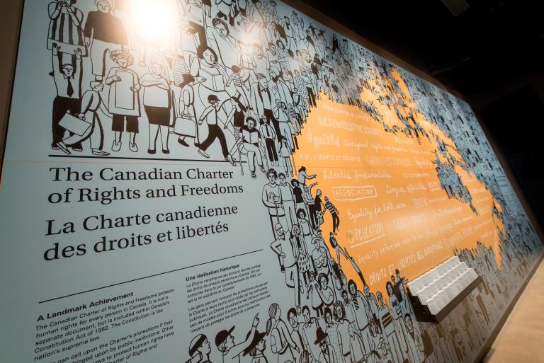 A museum exhibition wall features an illustrated mural of diverse figures and the title, “The Canadian Charter of Rights and Freedoms / La Charte canadienne des droits et libertés.” The mural transitions from black illustrations of people into an orange section with key rights and freedoms in both English and French in the shape of the Canadian Map. Partially obscured.