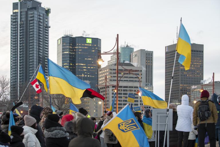 Une foule de personnes emmitouflées se tient debout dans une rue de ville pendant l’hiver, tenant de multiples drapeaux ukrainiens bleus et jaunes ainsi qu’un drapeau canadien. Le rassemblement semble être une manifestation en appui à l’Ukraine. De grands édifices à bureaux et des feux de circulation sont à l’arrière-plan sous un ciel couvert. Partially obscured.