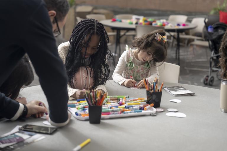 Two young children sit side by side at a round table, engaged in an art activity with markers and coloured pencils. Partially obscured.