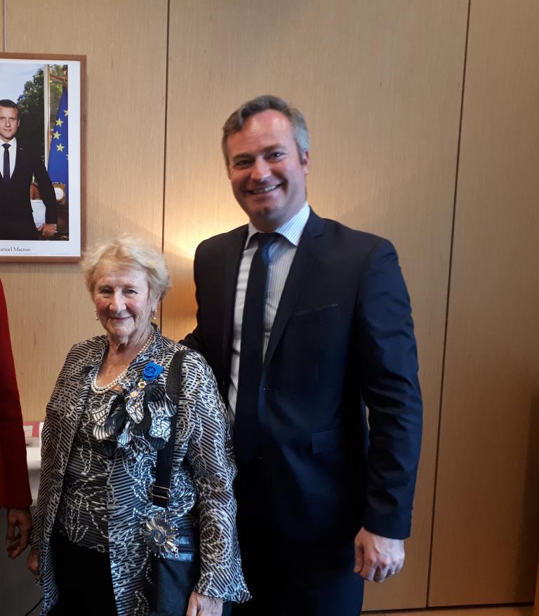 An elderly woman with short gray hair smiles gently at the camera. She is wearing pearl earrings, a pearl necklace, and a patterned black-and-white blouse. She stands in warm light in front of a wood-panelled wall. Partially obscured.