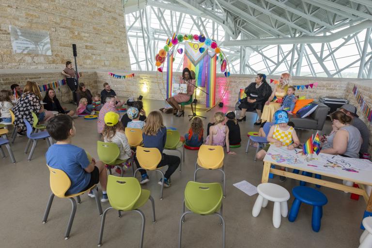 A drag artist reads aloud to a group of children and adults seated in colourful chairs inside a bright, industrial-style venue decorated with rainbow banners and bunting fans for a Pride-themed storytime event. Partially obscured.