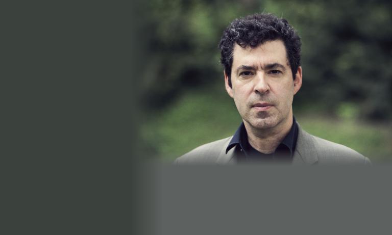 Headshot of a middle-aged man with dark curly hair wearing a grey blazer over a black shirt, photographed outdoors against a blurred green foliage background. His expression is serious and direct. Partially obscured.