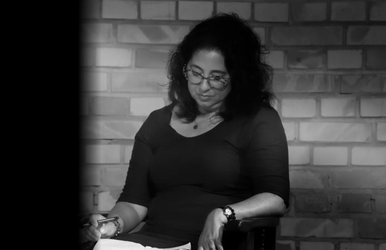 A black-and-white portrait of a woman with dark curly hair who is looking down at a paper on her lap. Partially obscured.