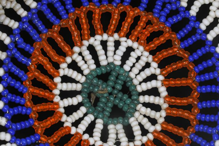 A close-up photo of a circular beadwork design on a black background. The pattern features concentric rings of turquoise, white, orange and bright blue beads arranged in a radiating floral or sunburst motif. Partially obscured.