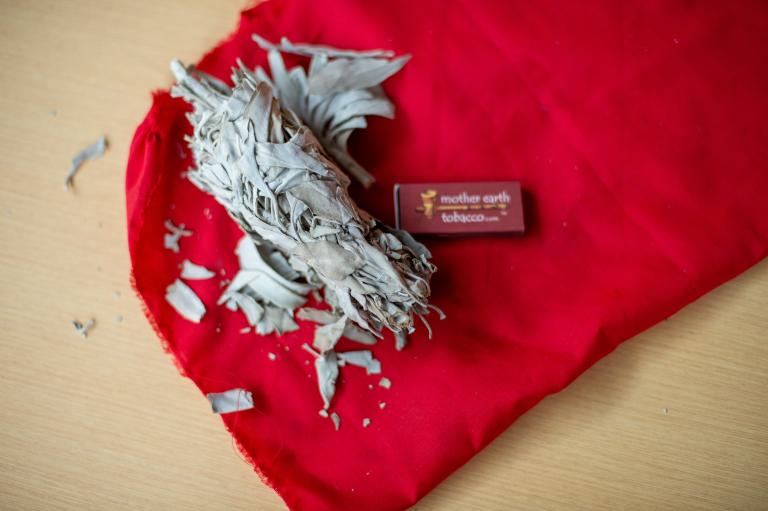 A small bundle of dried sage lies on a bright red cloth, with loose leaf fragments scattered around. Beside it is a small rectangular match box labeled “Mother Earth Tobacco.“ The items rest on a light wooden surface. Partially obscured.