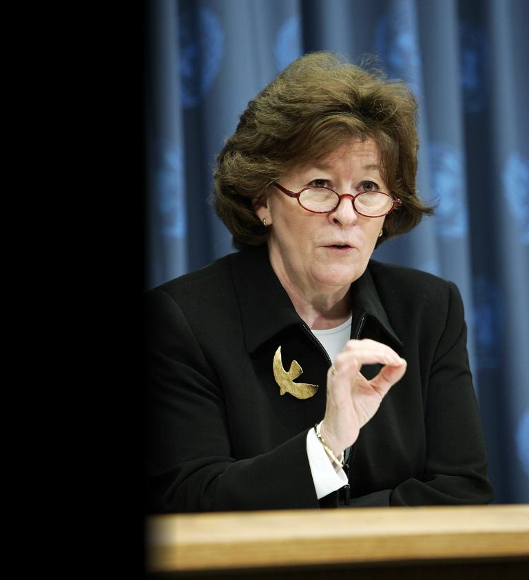 A woman with short brown hair and red glasses, wearing a black blazer with a gold bird brooch, is speaking at a podium while gesturing with one hand. Partially obscured.