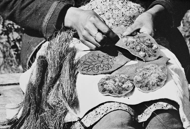 A black-and-white photo of hands holding and stitching decorated fabric pieces with floral embroidery and textured fibres spread across a person's lap. Partially obscured.