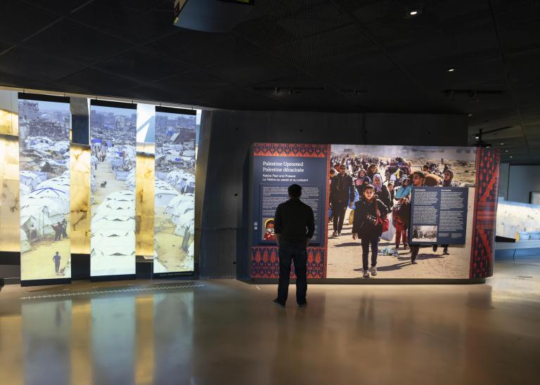 A wide shot of a Museum gallery. On the left side are three vertical panels showing a photo of a white tents and people displaced. On the right side of the image, a visitor is standing in front of an exhibit wall reading the text. Partially obscured.