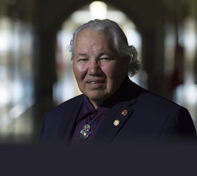 Portrait of an older Indigenous man with long gray hair pulled back, wearing a dark blazer, purple shirt, and decorative bolo tie, sitting in a softly lit hallway with an out-of-focus background. Partially obscured.