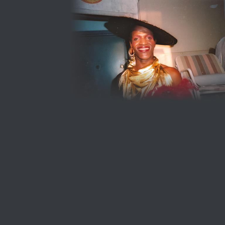 A smiling person dressed in glamorous performance attire, including a wide-brimmed black hat with embellished trim, a gold satin draped halter-style top, red feathered accessories and chandelier earrings. The photo appears to be a vintage snapshot taken in a backstage dressing room. Partially obscured.